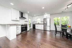 Bright, modern kitchen with white cabinets, stainless steel appliances, dark wood flooring, round dining table with chairs, and large windows letting in natural light.