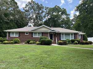 A single-story brick house at 211 Hillcrest Drive, Oxford, features light-colored trim, well-kept shrubs, and a large green lawn. A concrete walkway leads to the black front door, with tall trees encircling the property under a partly cloudy sky.
