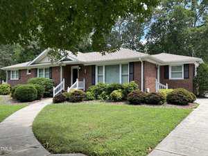 Single-story red brick house with white trim and shutters, a front porch with white railings, and a curved sidewalk. The house is surrounded by green bushes, trees, and a well-kept lawn.