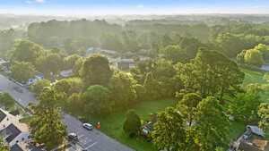 Aerial view of a residential neighborhood with tree-lined streets, houses, and a large grassy area. Morning light creates mist and shadows over the landscape, with dense woods in the background.