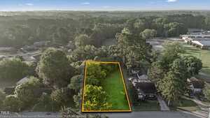 Aerial view of a vacant rectangular lot outlined in orange, surrounded by trees and neighboring houses, with a road on one side and a wooded area in the background.