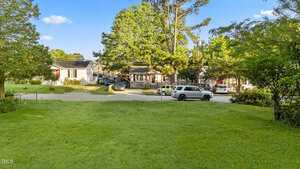 A grassy yard with a street and several parked cars in front of single-story houses surrounded by trees under a blue sky with scattered clouds.