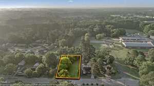 Aerial view of a residential neighborhood with a rectangular vacant lot outlined in orange near the center. The lot is grassy with several trees, and nearby houses and a large building are visible.
