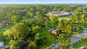 Aerial view of a suburban neighborhood with houses, green lawns, and dense trees; a large building complex, possibly a school, is visible among the trees, with roads winding through the area under a clear sky.