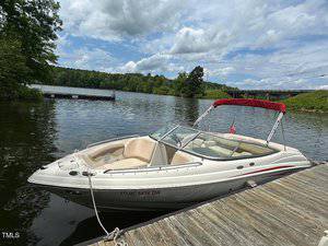 A white and beige speedboat with a red canopy is docked at a wooden pier on a lake. There is a small American flag on the boat. Trees and a dock are visible across the water under partly cloudy skies.
