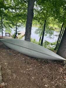 A gray canoe is turned upside down and resting on the ground between trees, with a view of a lake and a wooden dock in the background under a canopy of green leaves.