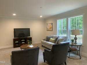 A living room with a beige sofa, two dark armchairs, a wooden coffee table, a TV on a stand, a floor lamp, and a window showing greenery outside. Neutral colors and simple decor create a cozy atmosphere.
