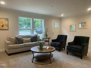 A living room with a light gray sofa, two dark armchairs, a round wooden coffee table, and neutral walls. Large windows show a green view outdoors, and a door is in the background. Artwork and mirrors decorate the walls.