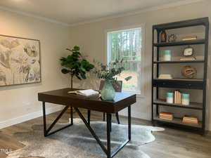 A modern home office with a dark wood desk, a green plant in a vase, a large wall painting, a tall bookshelf with books and decor, and a window showing trees outside. A gray rug is beneath the desk.