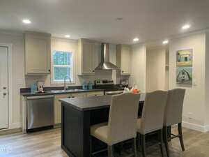 Modern kitchen with light tan cabinets, a stainless steel oven and dishwasher, a black countertop island with three beige upholstered chairs, and two paintings of houses on the wall. Wood flooring and recessed lighting are present.