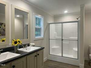 A modern bathroom with a double sink vanity, a vase of sunflowers, a window, a glass-enclosed shower, and a toilet. The room has light walls and wood-look flooring.