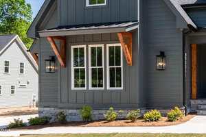 A close-up of a modern house exterior with gray vertical siding, a large white-framed window, wooden awnings, two black wall lanterns, stonework along the foundation, and landscaped shrubs in front.