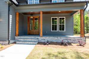 A modern house front porch with wooden columns, stone steps, gray siding, a wooden front door with glass panels, and two white-framed windows. Small plants are planted along the border of the porch.