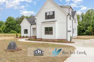 A modern white two-story house with black trim, surrounded by a lawn with fresh straw, minimal landscaping, and a curved driveway. Trees and a blue sky are in the background. Logos for ECI Construction and Hatcher Creek are visible.