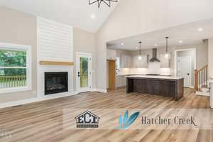 Spacious open-concept living area with light wood flooring, a modern fireplace, large windows, and a kitchen featuring white cabinets and a center island. Logos for ECI Construction and Hatcher Creek are overlaid on the image.