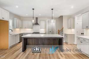 A modern kitchen with light gray cabinets, stainless steel appliances, a large dark island with pendant lights above, wood-style flooring, and white subway tile backsplash. Logo overlays at bottom center and right.
