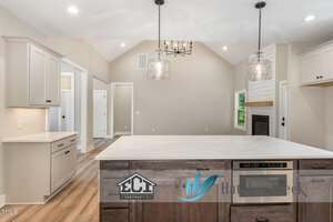Modern kitchen with light-colored cabinets, marble island countertop, and wooden flooring. Two pendant lights hang from the ceiling. The adjoining living area features a fireplace and large windows. Logos are visible on the island.