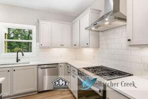 A modern kitchen with white cabinets, stainless steel appliances, a gas stove, white subway tile backsplash, a window above the sink, and logos for ECI Construction and Hatcher Creek on the image.