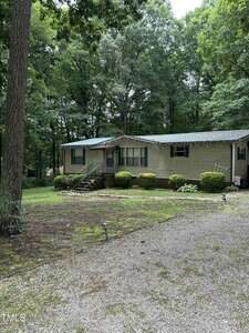 A single-story beige manufactured home at 85 Southside Estates Court in Henderson sits among tall trees. There are metal stairs leading to the front porch, bushes along the house, and a gravel driveway in the foreground.