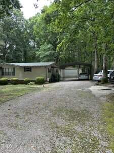 A single-story beige house with green trim sits among trees. A gravel driveway leads to a carport with two cars parked underneath. Bushes line the house, and a wooded area surrounds the property.