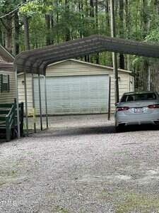 A light gray car is parked under a metal carport on a gravel driveway in front of a beige garage with a closed white door, surrounded by tall trees and greenery.
