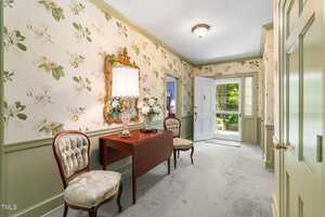 A hallway with floral wallpaper, light green wainscoting, a wooden console table with a lamp and flowers, two upholstered chairs, light carpet, and a door open to a green yard outside.