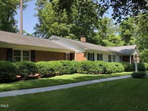 Single-story brick house with white trim and black shutters, surrounded by trimmed green bushes and trees. A concrete walkway runs along the front, and the lawn is well-maintained under a clear sky.