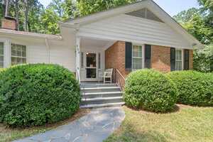 A brick and white siding house with black shutters, a small front porch, two white chairs, gray steps, and large green bushes bordering a stone pathway.