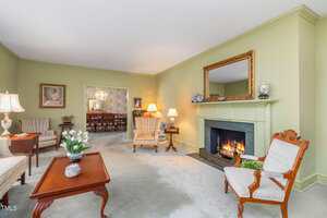 A traditional living room with light green walls, carpeted floor, a fireplace with a mirror above it, several armchairs, side tables with lamps, and decorative items. A dining room is visible through a wide doorway.