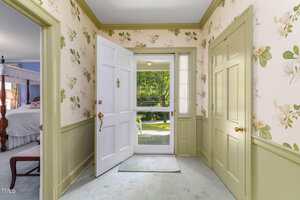 A hallway with floral wallpaper, green trim, and light carpet. The front door is open, revealing a view of a green lawn and trees outside. Two closed doors are on the right, and a bedroom is visible to the left.
