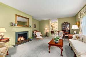 A traditional living room with green walls, a fireplace with a mirror above it, classic wooden furniture, a coffee table with flowers, armchairs, a sofa, lamps, and a china cabinet in the background.