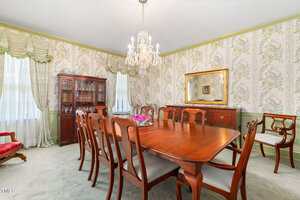 A formal dining room with a wooden table and eight chairs, a chandelier, a china cabinet, a sideboard, floral wallpaper, carpeted floor, and two windows with drapes. A large mirror hangs on one wall.