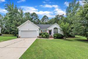Charming single-story white house in Creedmoor at 2003 Zbonack Court, with a two-car garage, front steps leading to the entrance, neatly trimmed bushes, and a large grassy lawn surrounded by trees under a partly cloudy sky.