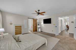 A bedroom with a ceiling fan, a bed with light bedding, a dresser, a wall-mounted TV, and a sliding barn door leading to a bathroom. The room has beige walls and carpeted flooring.