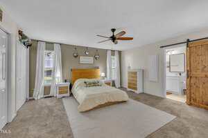 A bedroom with a large bed centered against a wall, two windows with white curtains, matching wooden nightstands, a ceiling fan, beige carpet, a barn-style sliding door, and a view into an adjacent bathroom.