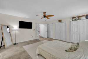 A bedroom with beige carpet, a bed with a light-colored comforter, a wall-mounted TV, a standing mirror, a floor lamp, multiple white closet doors, and a ceiling fan with wooden blades.