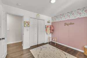 A children's room with floral wallpaper and pink paneling on one wall, a name sign reading "Brynlee," hanging clothes, a basket, a decorative rug, and double white closet doors.