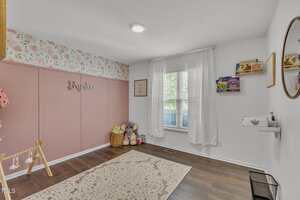A child's bedroom with pink and white walls, a floral wallpaper border, a name sign reading "Brynlee," a window with white curtains, a rug, toys, and shelves with books and decor.
