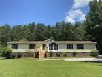 Single-story manufactured home at 4037 Crestview Lane in Oxford features a brick foundation, beige siding, and brown roof. Wooden steps lead to the front door, with a grassy lawn, shrubs, and trees under a partly cloudy blue sky.