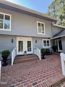 A two-story gray house with white trim features double front doors, brick steps, and a large brick porch. Potted plants and a black bench are near the entrance, and trees are visible in the background.