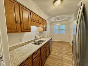Galley kitchen with wooden cabinets, granite-style countertops, double sink, stainless steel appliances, light wood flooring, white walls, and a small dining area with windows at the far end.