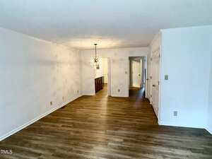 A mostly empty room with white walls, wood-style flooring, a light fixture hanging over the dining area, and a view into a kitchen and hallway. Double white closet doors are visible on the right.