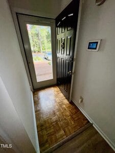 A small entryway with a parquet wood floor, a partially open black front door, a wall-mounted control panel, and a glass storm door leading outside to a brick pathway and green yard.