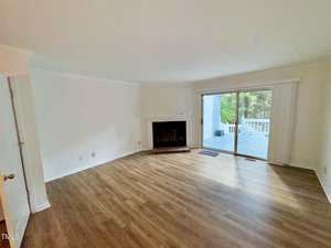 A spacious, unfurnished living room with wood flooring, a fireplace, white walls, and large sliding glass doors leading to a deck with a view of greenery outside.