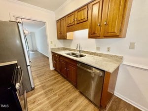 A galley kitchen with wood cabinets, light granite countertops, a double sink, stainless steel dishwasher, and refrigerator. The floor has light wood-style vinyl, and the walls are white with crown molding.