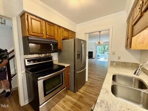 A kitchen with stainless steel appliances, including a stove, microwave, and refrigerator, wood cabinets, granite countertops, a double sink, and a view into a living area with wood flooring and a chandelier.