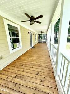 A long wooden front porch with white railings, two ceiling fans, beige siding, three windows, a gray door, and outdoor lights, overlooking a grassy yard with trees in the background.