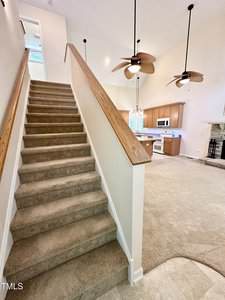 Carpeted staircase with wooden handrails leading up, next to an open living area featuring a kitchen with wooden cabinets, white appliances, ceiling fans, and a fireplace.
