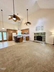 Spacious open-concept living area with beige carpet, a stone fireplace, ceiling fans, and a kitchen featuring wood cabinets, an island, and white appliances. Three framed artworks hang above the fireplace.