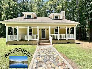 A tan house in Manson with a large front porch, white railings, and dormer windows sits in a grassy yard bordered by trees at 15 Wild Fox. An inset shows a lake with "WATERFRONT" in bold black letters.